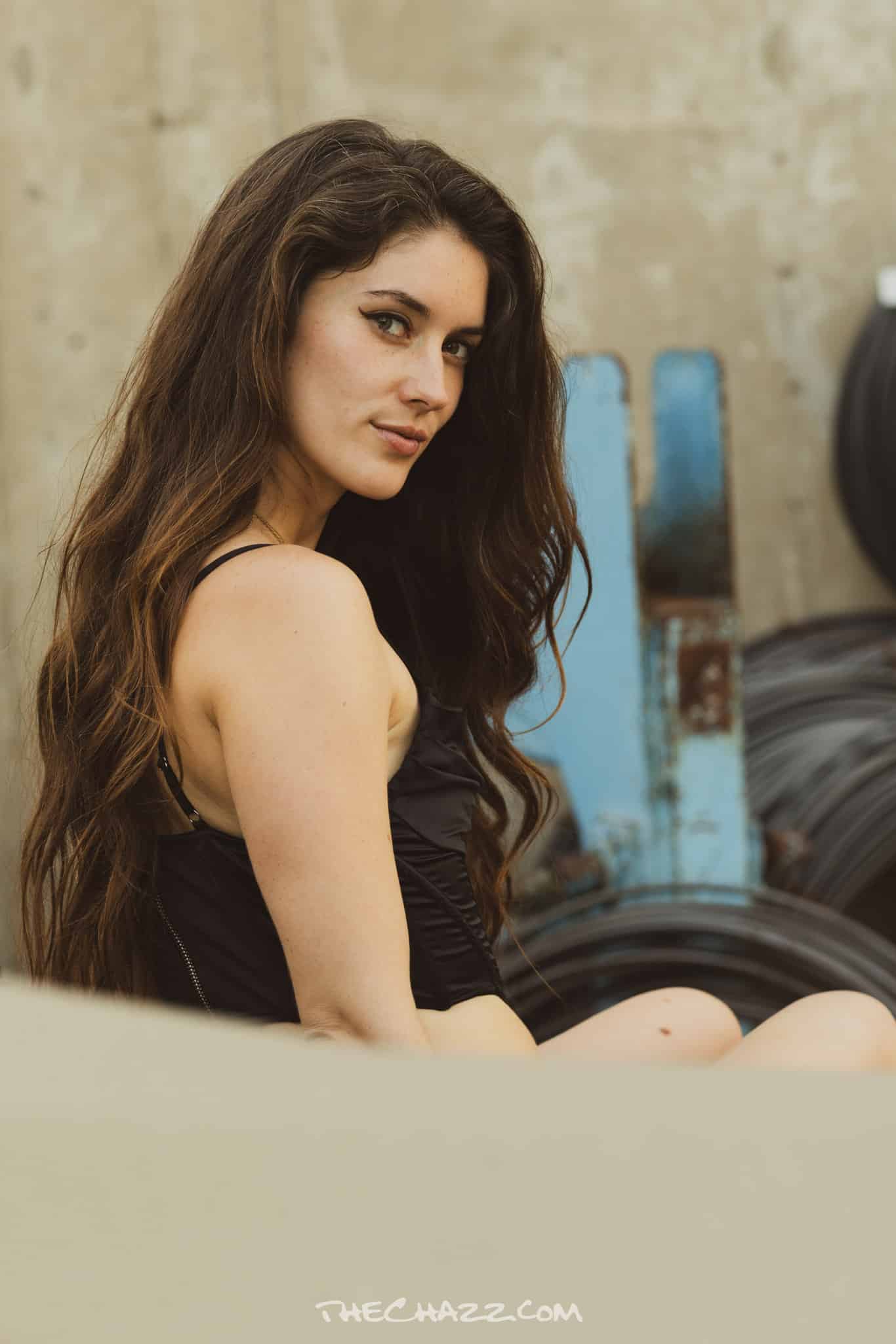 Beautiful young woman with long wavy brown hair at a gym, sitting against a wall of tires.